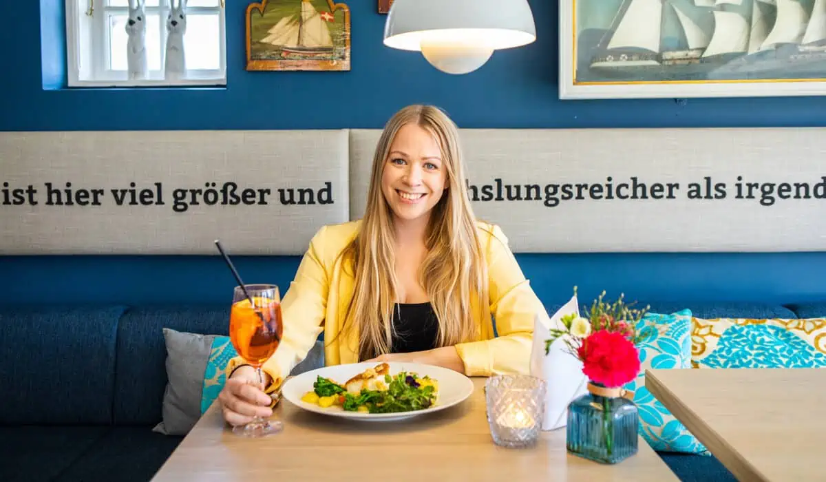 Kaamp Meren Kampen: Finja am Tisch im Innenraum mit Getränk in der Hand und Essen vor sich